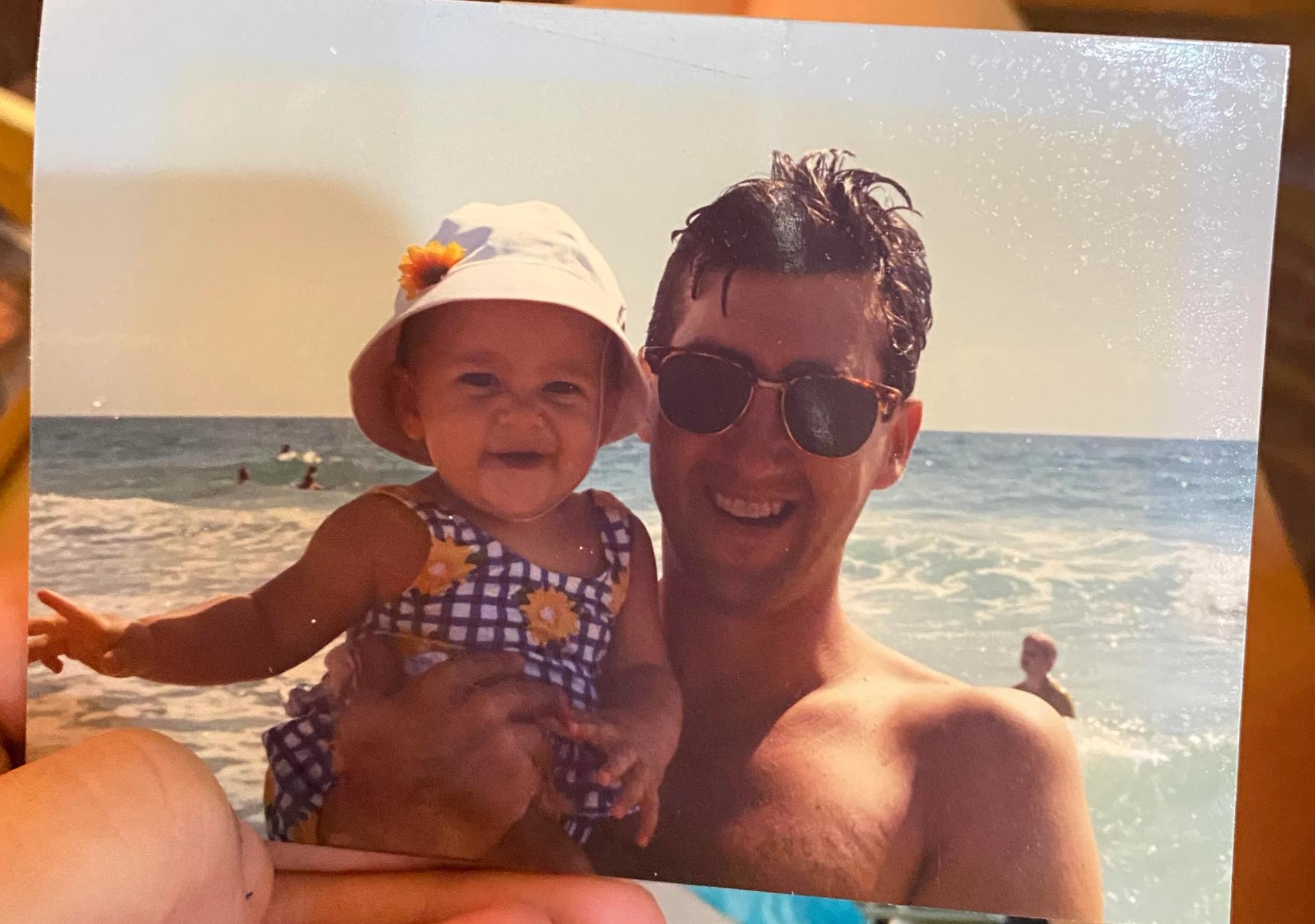 A baby wearing a white bucket hate and a black and white gingham swimsuit being held by her dad, who is wearing sunglasses, at the beach.