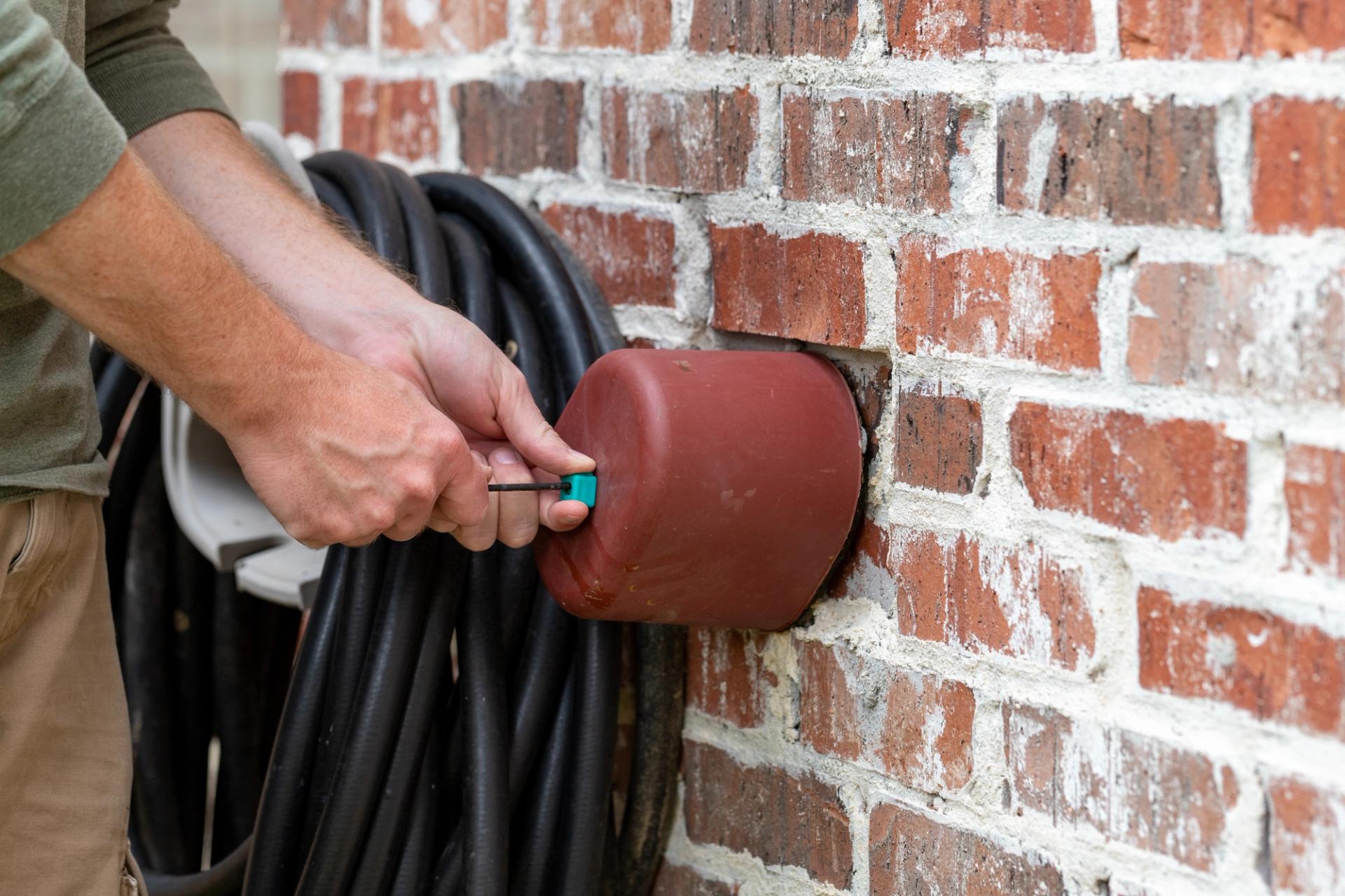Two hands puts a brown cover on an outdoor faucet on a red brick wall.