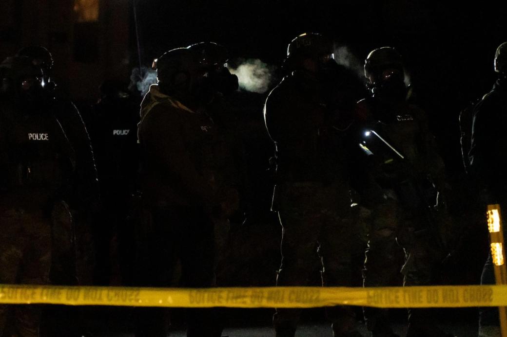Several federal agents outfitted with helmets, body armor, and firearms stand silhouetted in the dark behind a yellow "POLICE LINE DO NOT CROSS."