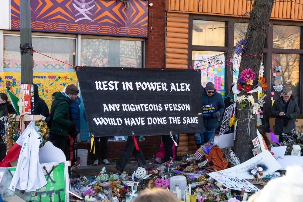A sign reading "Rest in power Alex. Any righteous person would have done the same" hangs on a mural-covered building, surrounded by flowers, candles, and other memorial items with mourners gathered nearby.