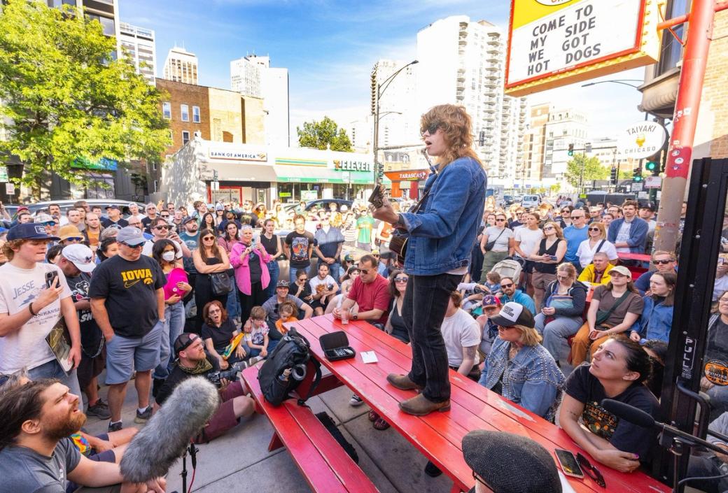 A man standing on a red table. he is holding a guitar and singing. crowded around him is a group of people watching him perform