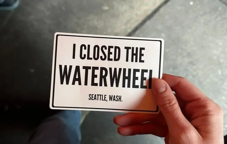 A hand holding a sticker with black text on a white background reading “I closed the waterwheel Seattle, Wash.”