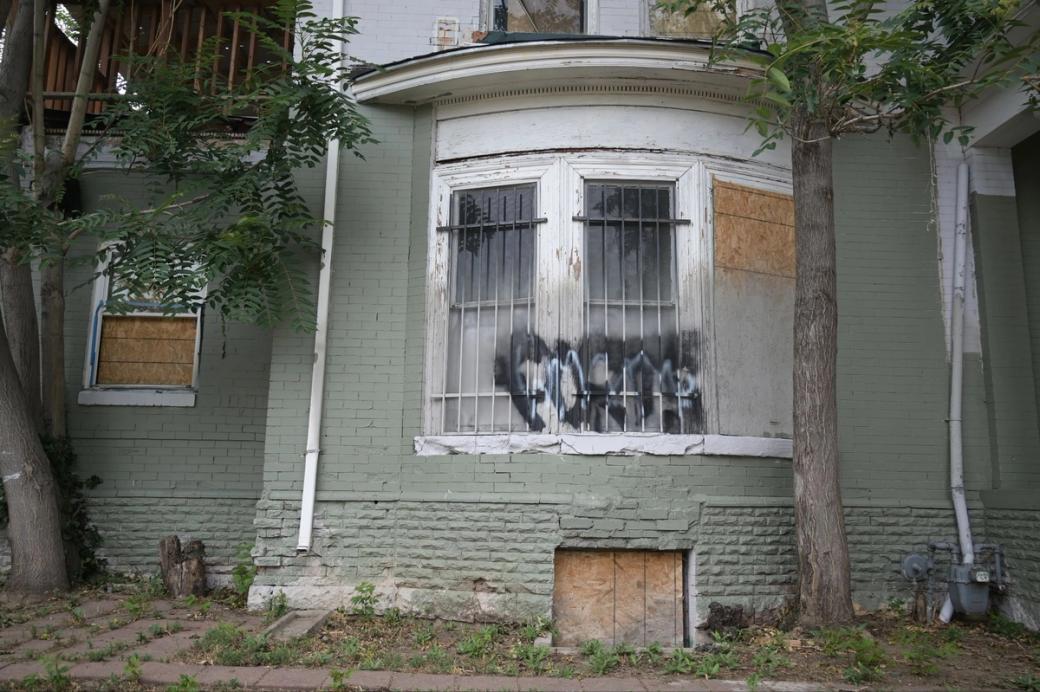 A light green building with white trim. The paint is fading in places, there is graffiti on some of the windows, and others are boarded up.
