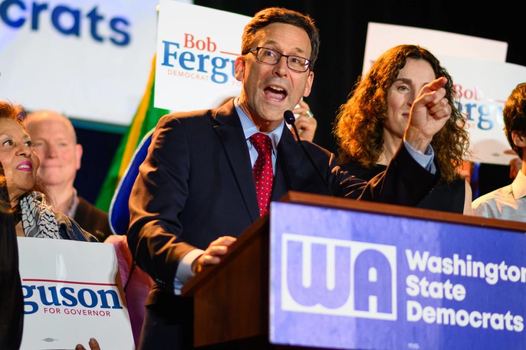 A man at a podium on a stage surrounded by people holding signs that say Bob Ferguson