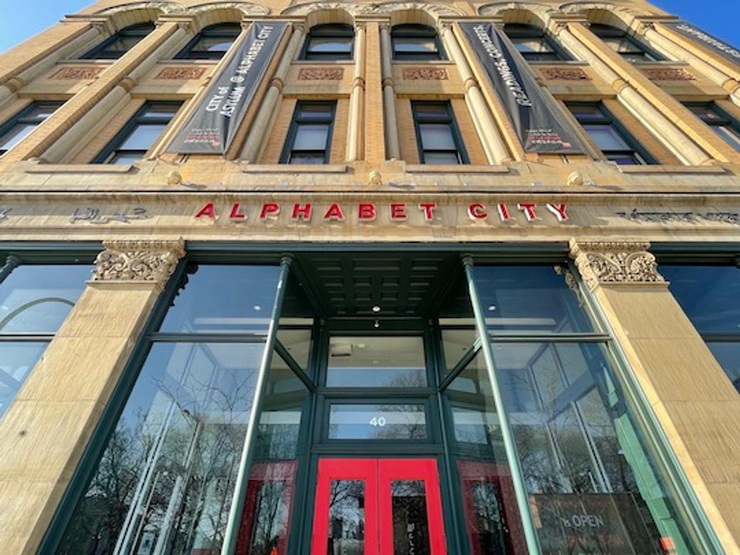 A tan victorian-era building with a red door and red letters reading "Alphabet City"