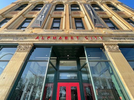 A tan victorian-era building with a red door and red letters reading "Alphabet City"