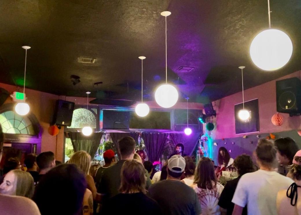 A crowded room of people watching musicians play on a stage with lights hanging from the ceiling.