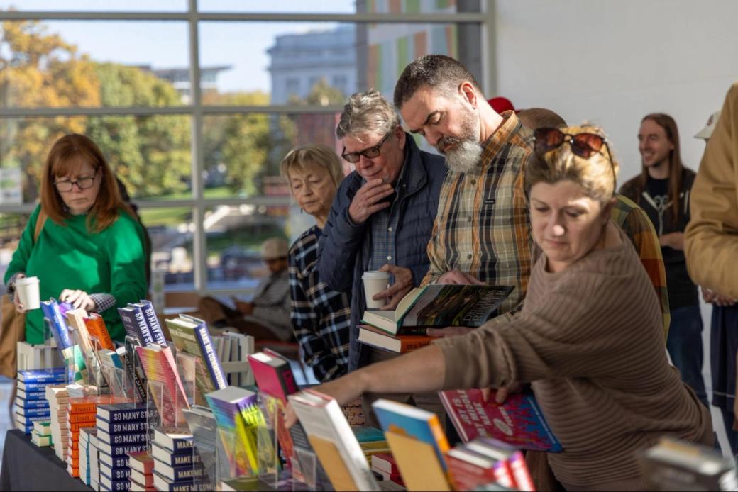 A group of people browse a table of books in a bright, sunlit space.