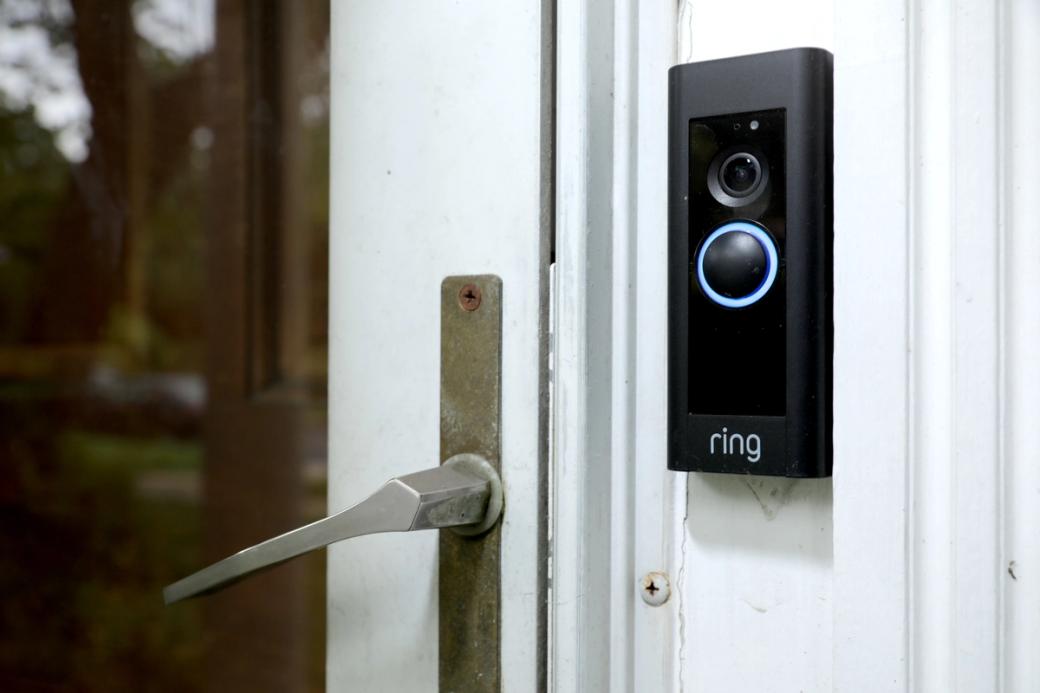 A black rectangular Ring doorbell device is affixed to a doorframe. The doorbell is lit up in light blue. 