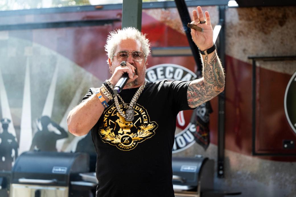 Guy Fieri stands on a stage holding a mic in one hand and raising his hand above his head with a smoking cigar pinched between his fingers. He has his classic bleached blond and spiked hair, sunglasses and two gold chains around his neck.