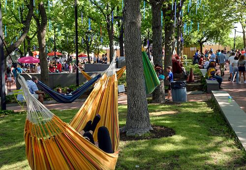 A person lounges in an orange hammock strung between two trees in a park, with dozens of people in hammocks, walking, and hanging out in the background.