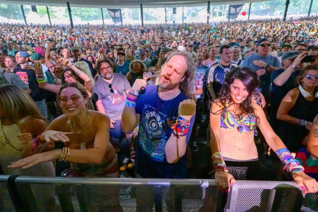 A crowd wearing colorful shirts and bracelets smiles and looks on at a Phish concert.