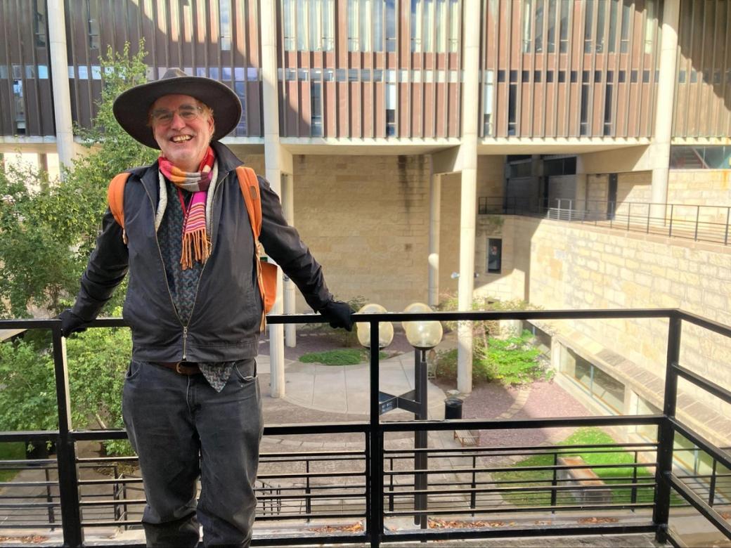 Madison artist Doug E.L. Haynes, dressed in black, leans against a black metal railing in front of the Mosse Humanities Building at UW Madison, a light-colored brick building.