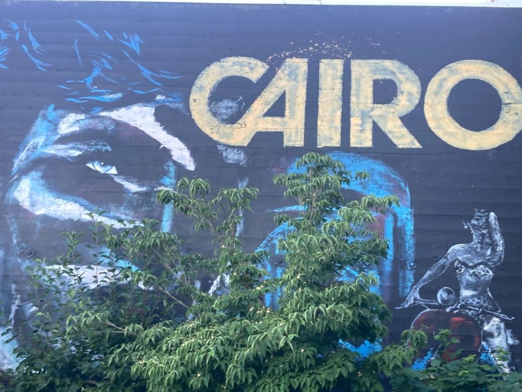 A large blue and white mural of a face, with a woman on a motorcycle in the bottom right corner and the word "CAIRO" in large yellow capital letters in the top right corner. 