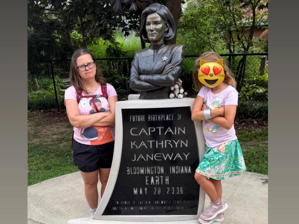 Two people stand with crossed arms on either side of a bust statue and plaque marking the "Future Birthplace of Captain Kathryn Janeway," Bloomington, Indiana, Earth, May 20, 2336, both mimicking the statue's stern, arms-crossed pose.