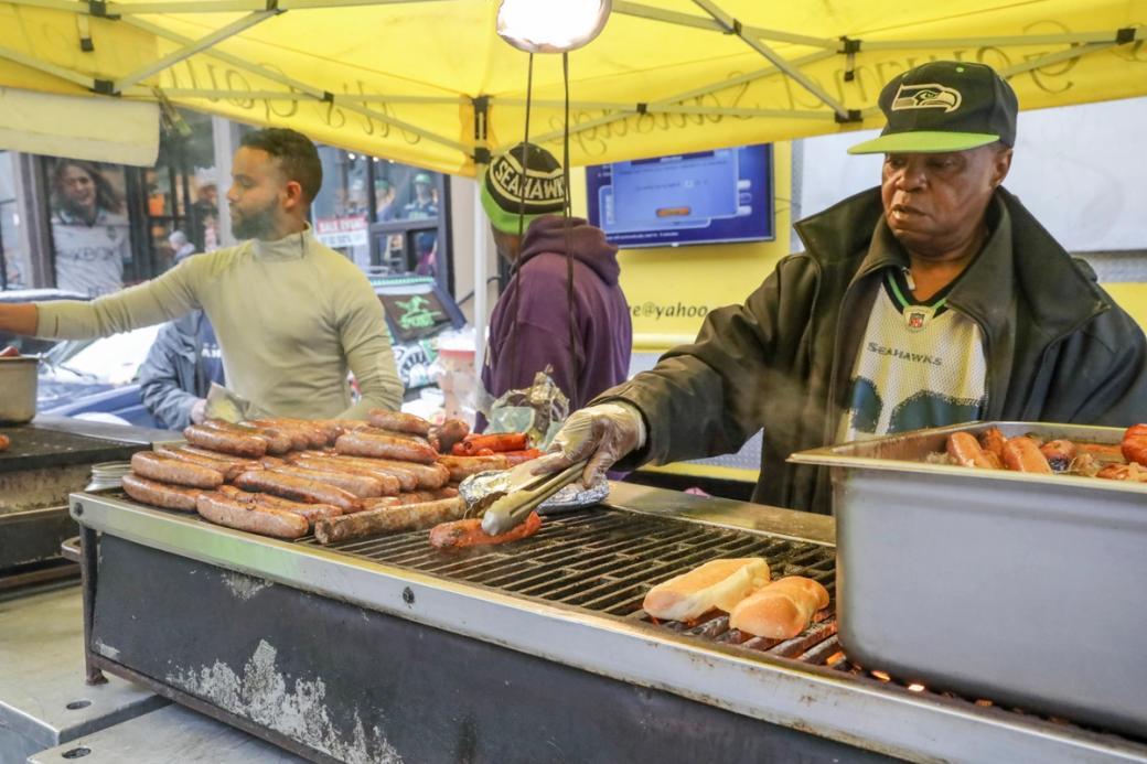 Licensed hot dog vendors wearing Seahawks gear grill up a pile of hot dogs under a yellow tent.