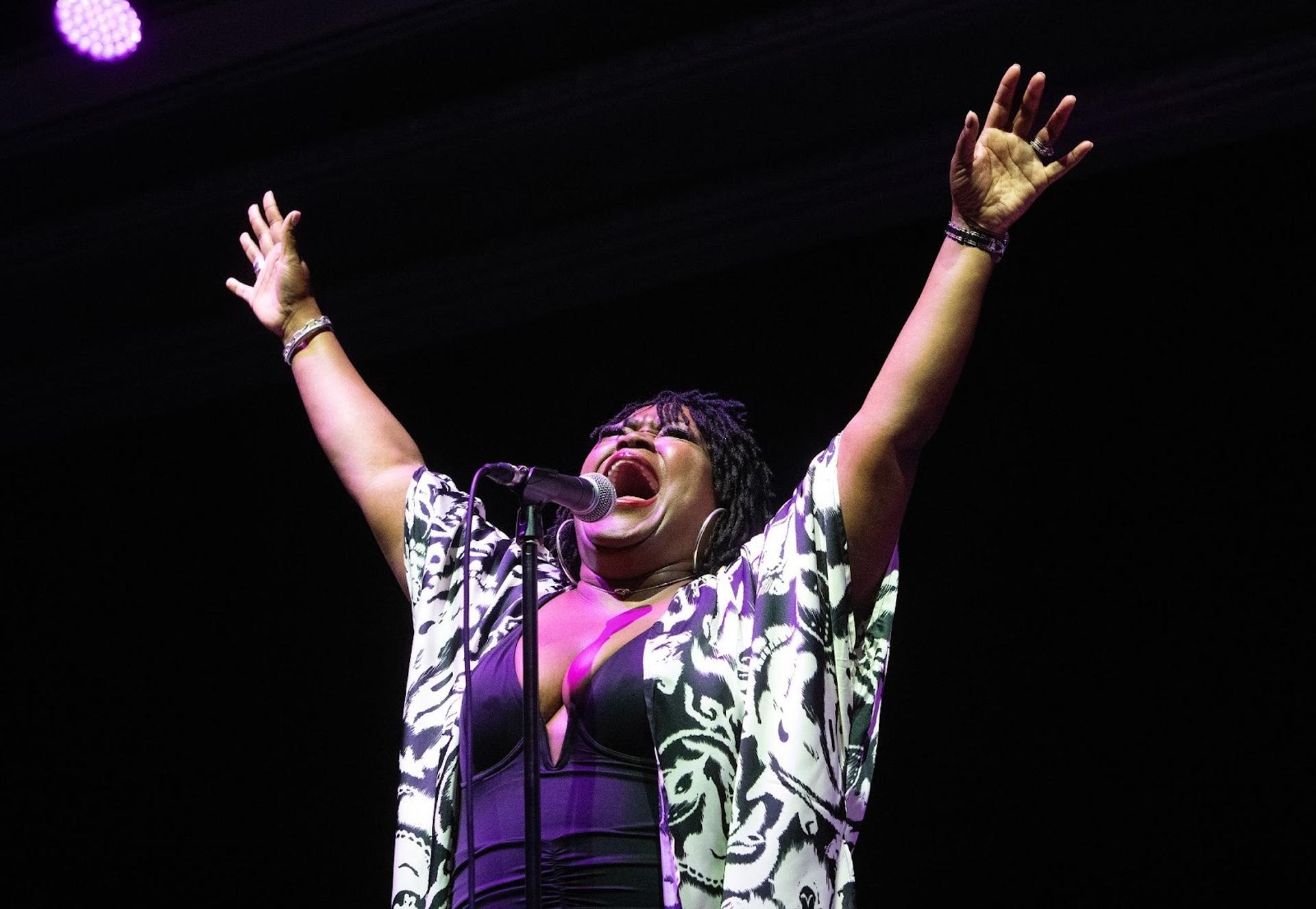 A woman sings with all her might into a microphone with both her arms in the air.
