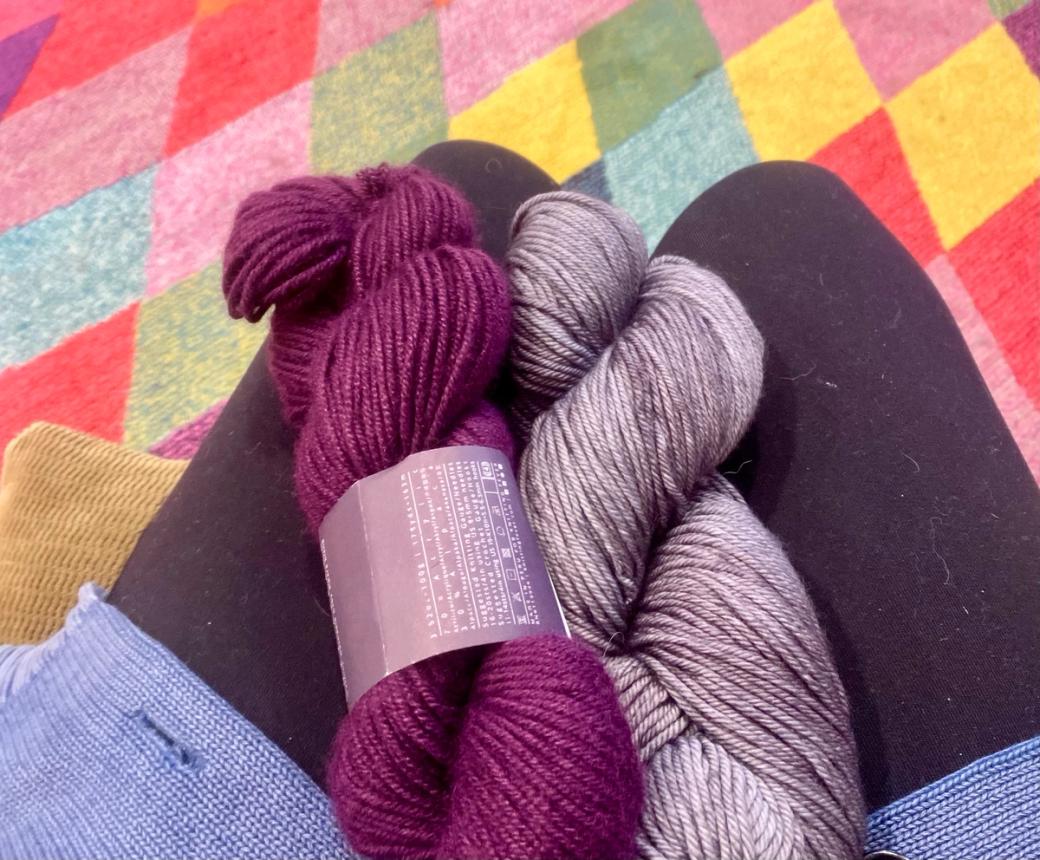 My lap with two skeins of yarn (one purple, the other grey) on it. The colorful floor of McWalker is in the background.