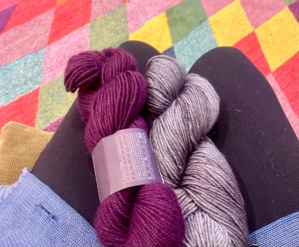 My lap with two skeins of yarn (one purple, the other grey) on it. The colorful floor of McWalker is in the background.
