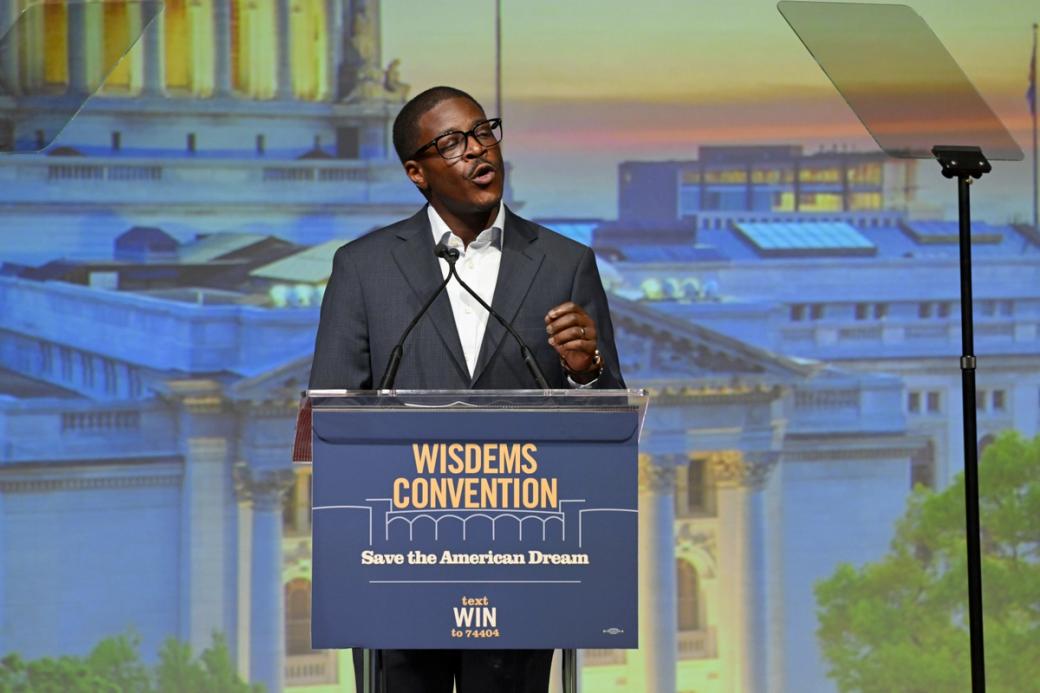 A man stands at a podium in front of a blue backdrop. 