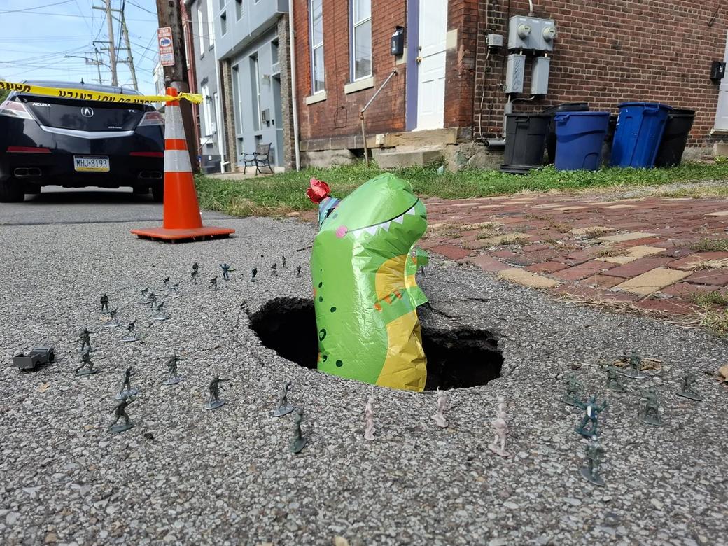 A lime green mylar dinosaur balloon sits in a small sinkhole and is surrounded by toy soldier army men.
