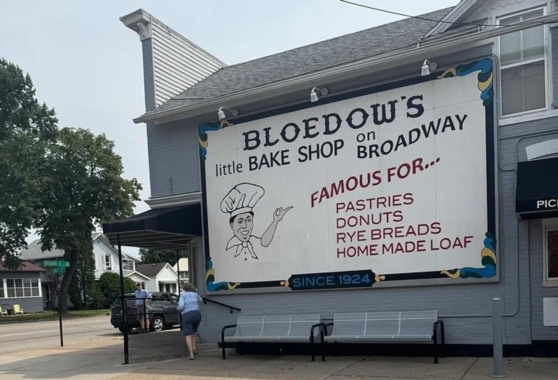 A building with a big sign reading "Bloedow's litte bake shop on broadway. Famous for pastries donuts rye breads home made loaf"
