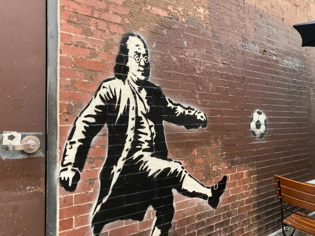 Black-and-white street art on a brick wall depicting Benjamin Franklin kicking a soccer ball.