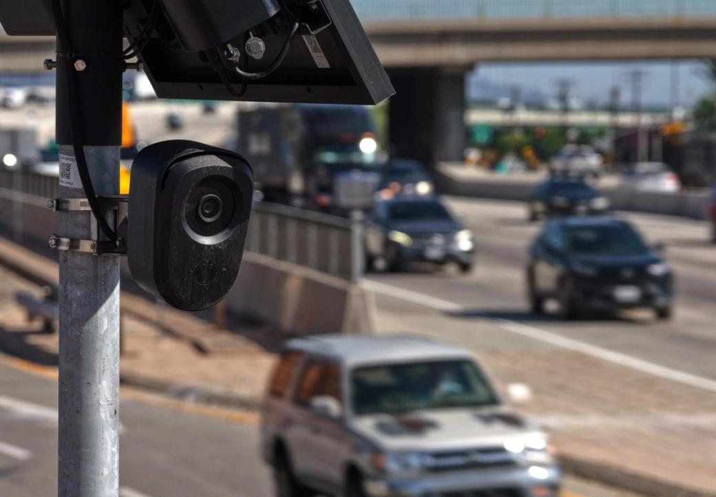 A camera mounted on a metal pole overlooks a busy multi-lane roadway, with several cars traveling beneath an elevated bridge in the background.