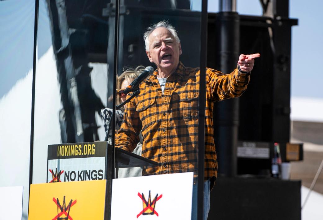 Adult man in plaid button up shirt speaks at podium with arm outstretched and hand pointing