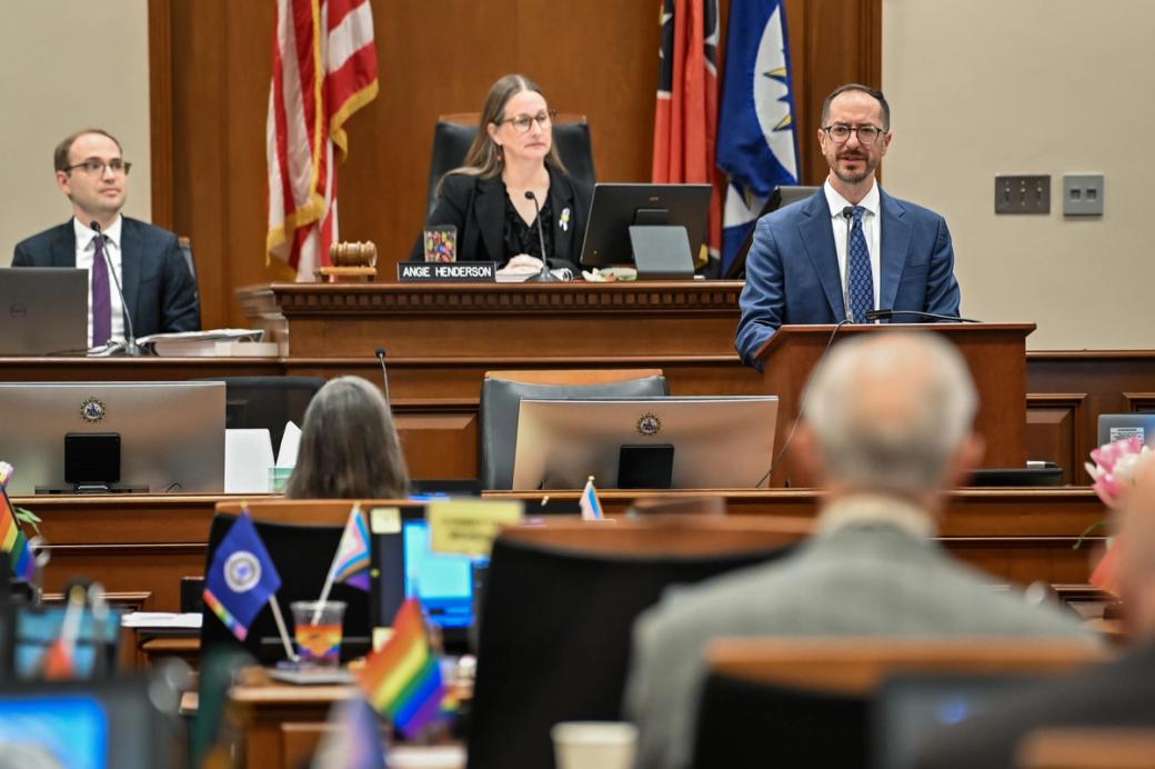 A white woman, Vice Mayor Angie Henderson, sits at a wooden judge bench, while a white man, Mayor O'Connell, speaks to councilmembers. We see the backs of councilmember heads as they sit at computers.