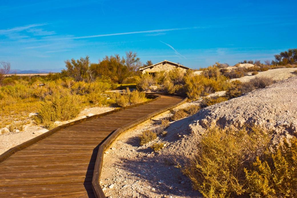 A wooden trail in the Ash Meadows National Wildlife Refuge leading to a small stone cabin surrounded by desert plants.