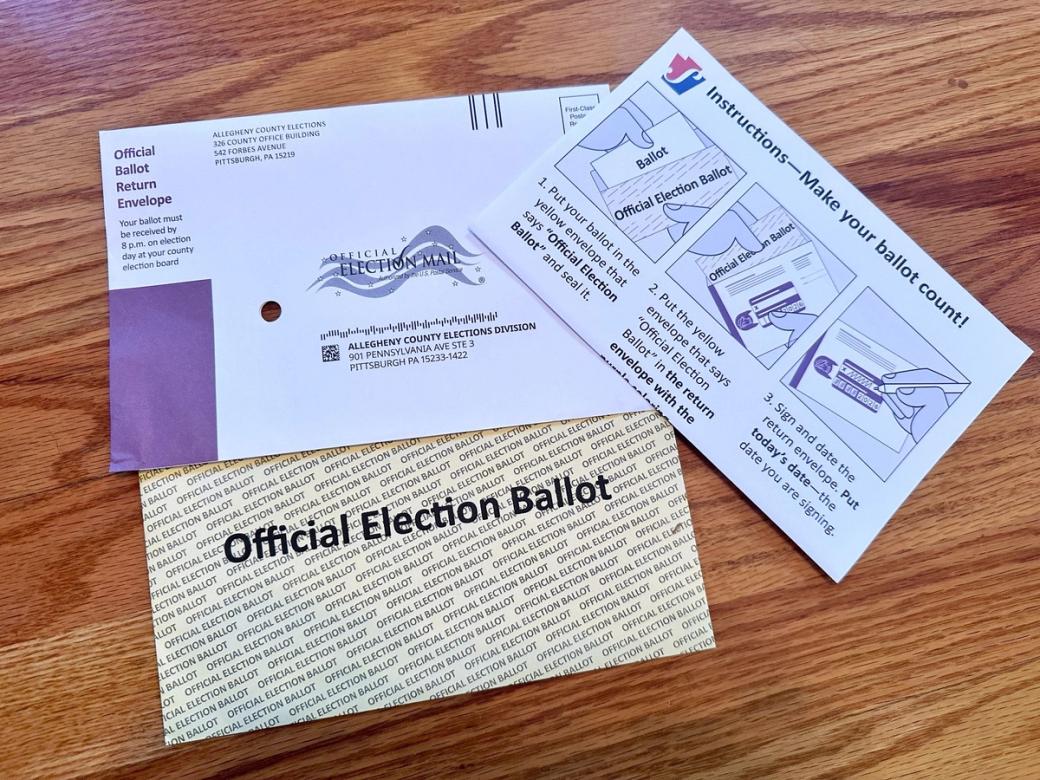 Three slips of paper — an official Allegheny County ballot return envelope, a yellow secrecy envelope, and a set of instructions — arranged on a hardwood floor.