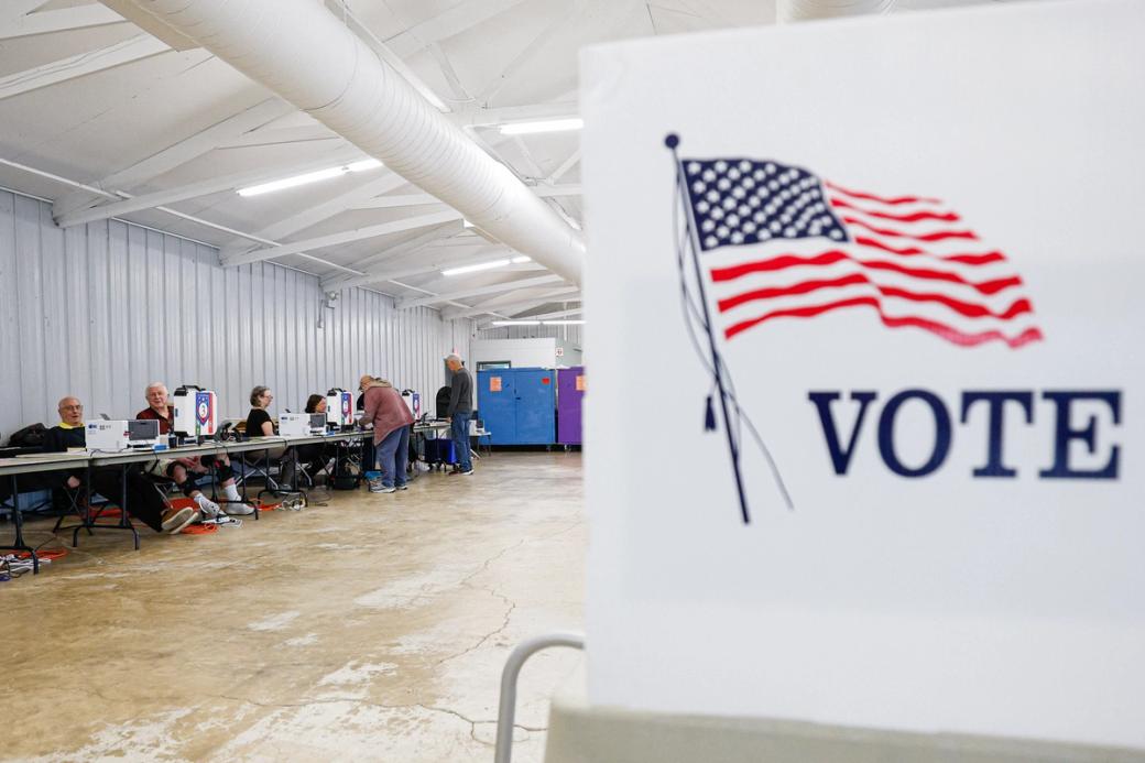 A polling place with election workers seated at tables and voters using voting equipment in a large indoor space. A large sign in the foreground displays an American flag and the word “VOTE.”