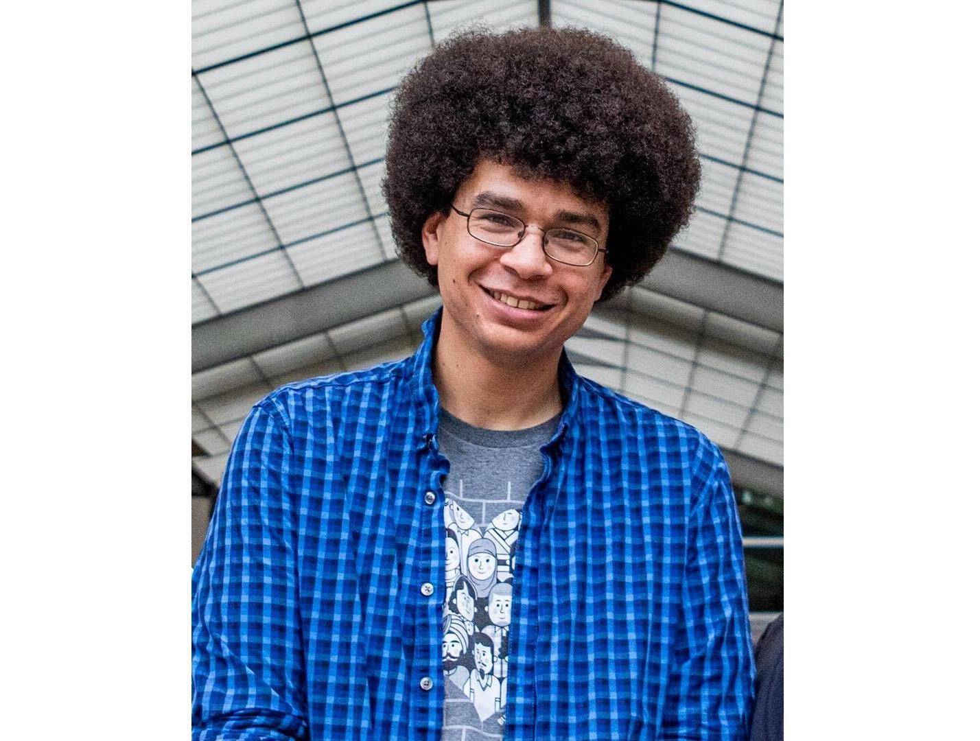 A man with brown, voluminous hair wearing a blue and white checkered shirt with a gray graphic T-shirt on underneath. The man is smiling.