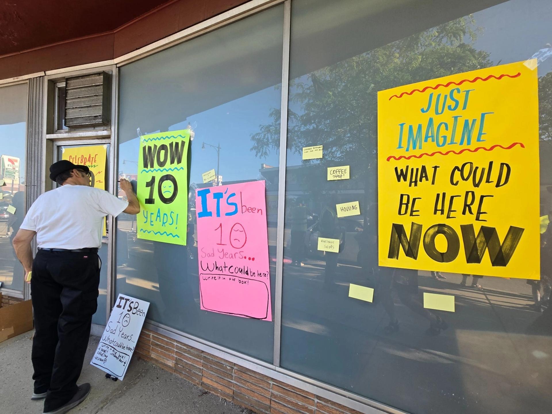 A man writes on a poster on the building. Posters read "Just Imagine what could be here now" post its are also placed on the window