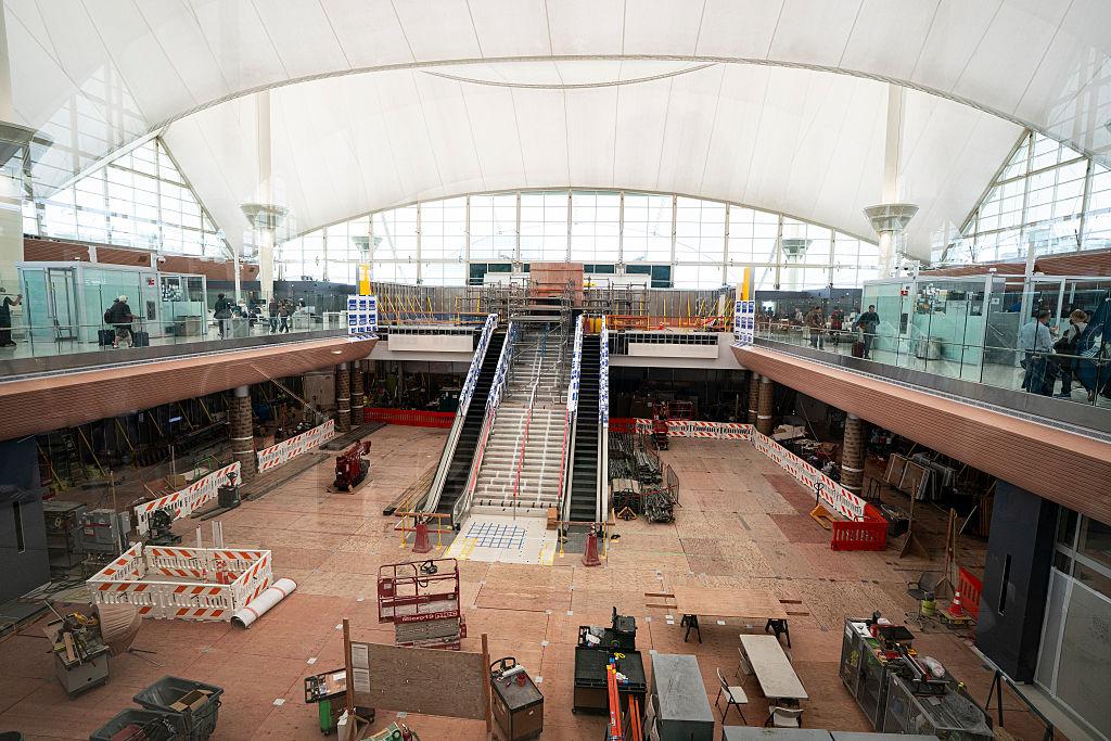 The interior of Denver International Airport's Great Hall under active renovation, with construction equipment, scaffolding, and orange barriers scattered across the floor beneath the building's distinctive white arched roof.