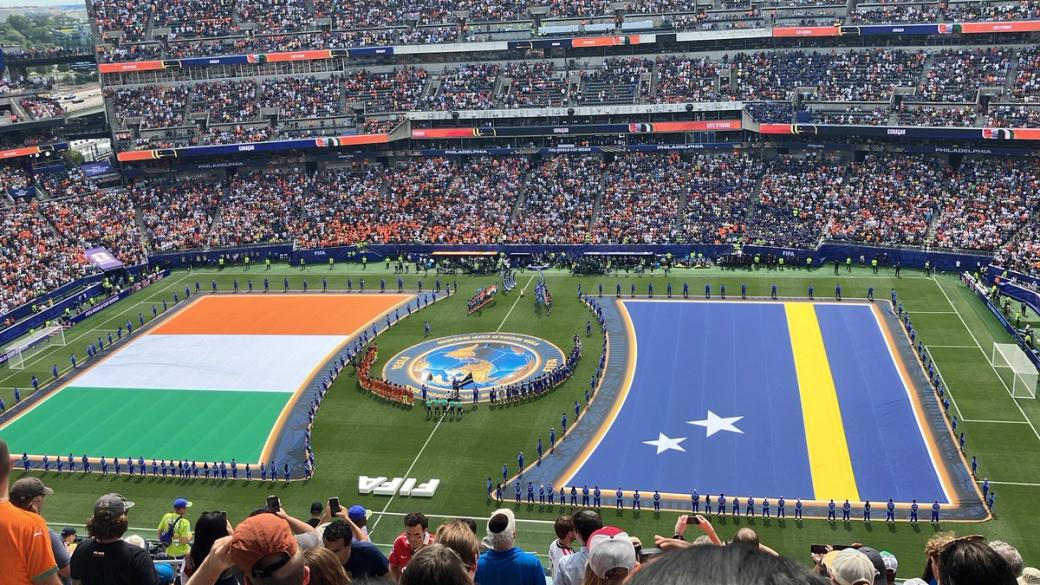 A tricolor flag of orange, white, and orange and a blue flag with two white stars and a yellow stripe are held up on a soccer pitch in a stadium