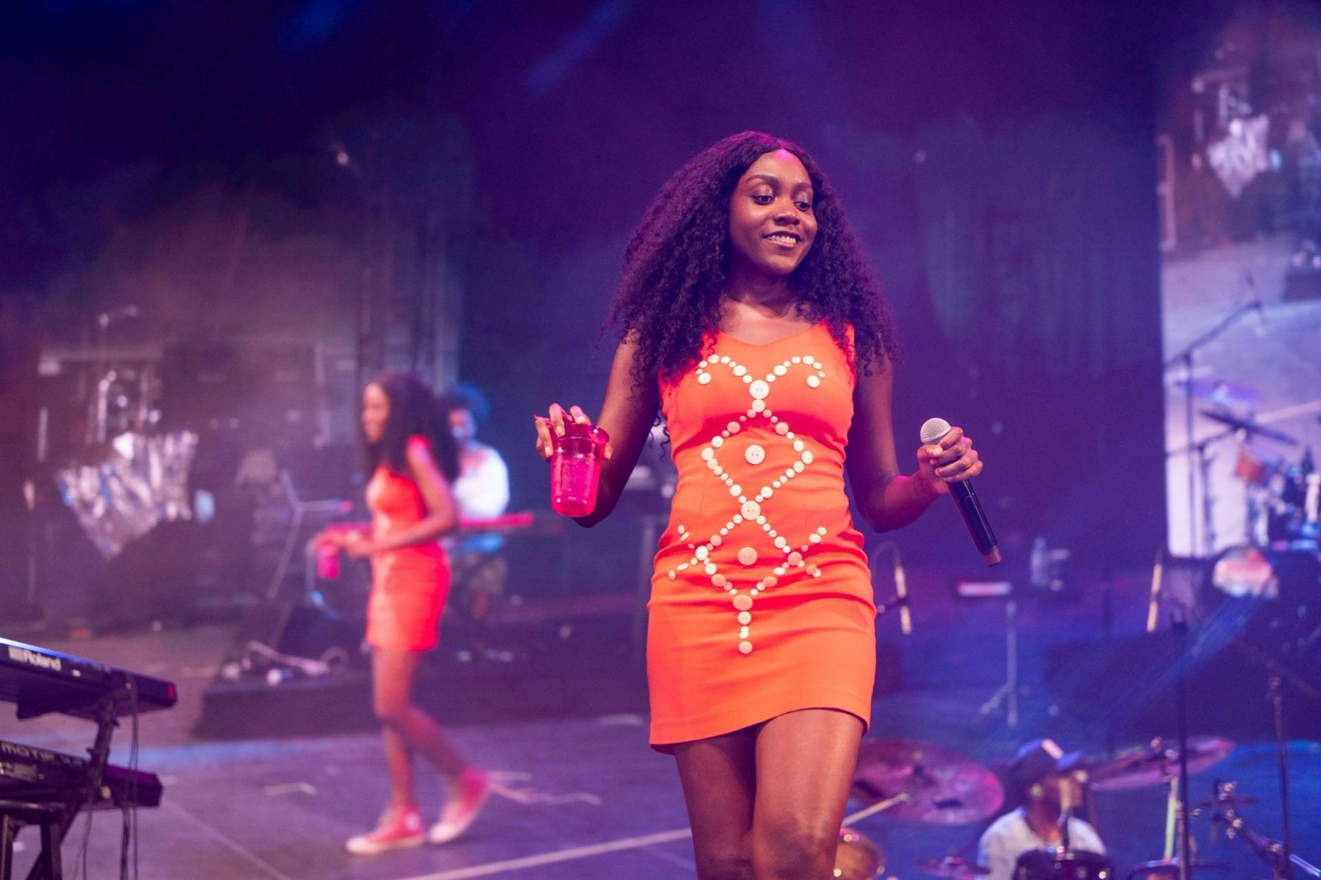 A woman in an orange dress holding a cup and a microphone. In the background it shows a screen from a side angle of this person