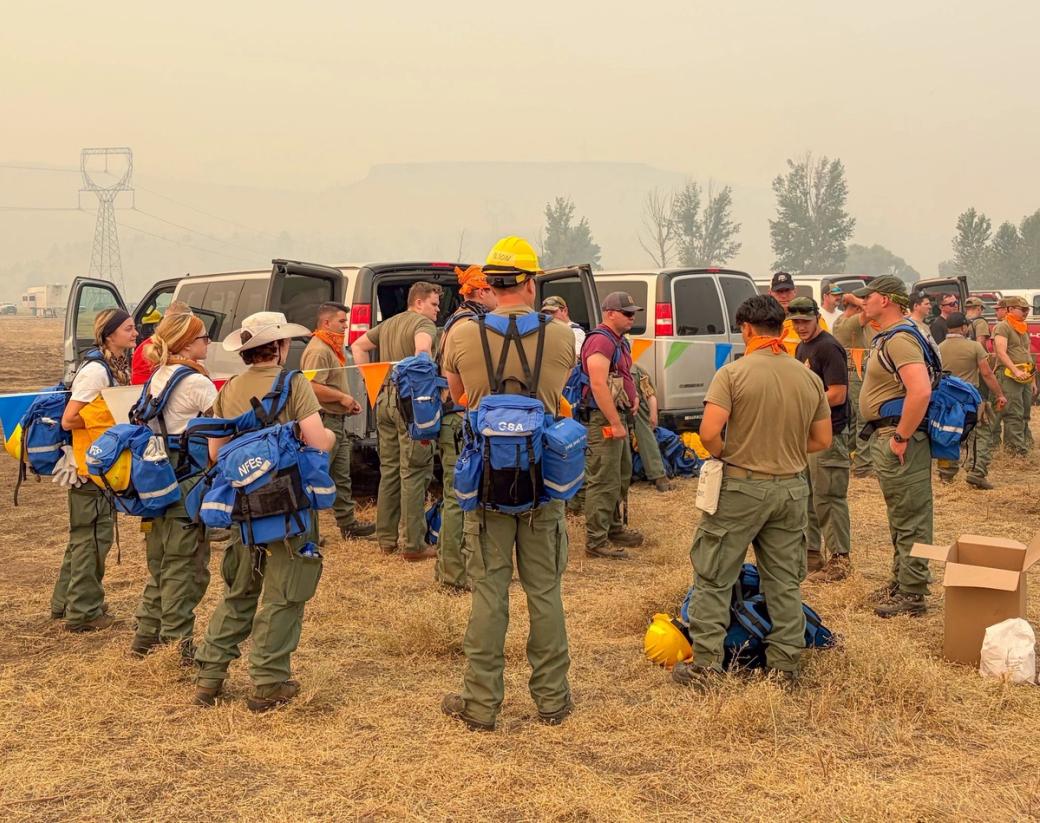 National Guard in green pants with blue backpacks arrive in a dry grass field, vans in the background