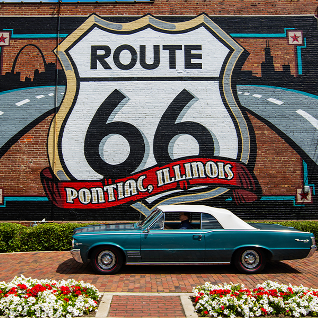 Display Ad: A teal vintage car is parked in front of a vibrant Route 66 mural in Pontiac, Illinois. The mural features bold lettering and a road graphic, creating a nostalgic atmosphere.