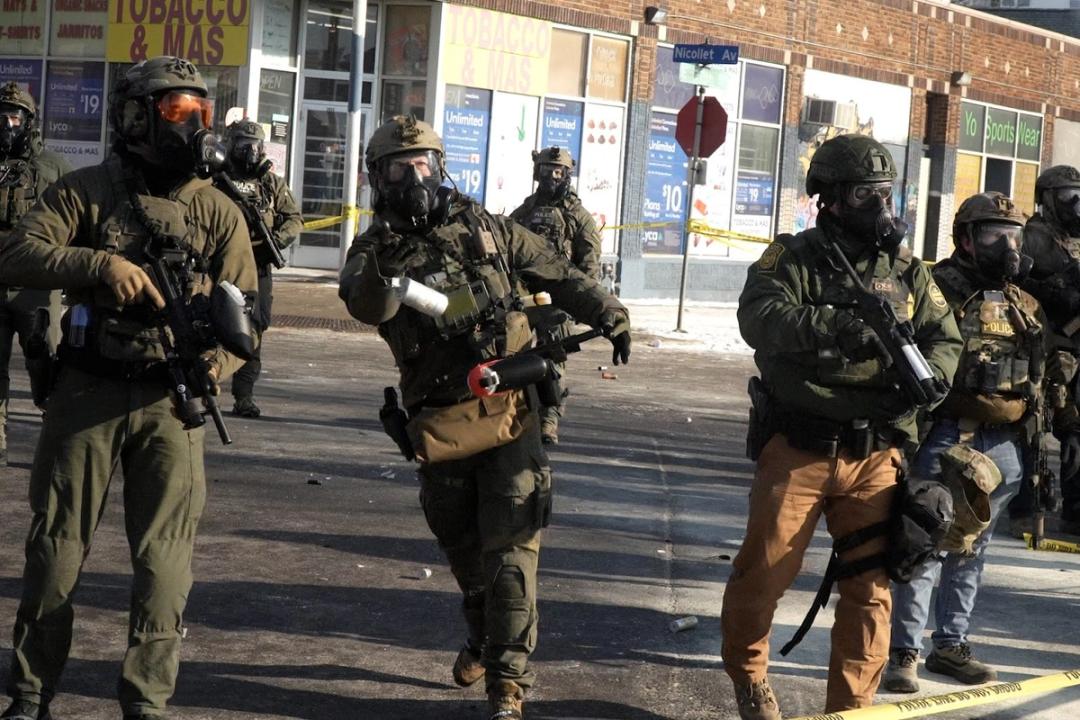 A federal officer in full tactical gear is throwing a tear gas canister toward the camera. Several other officers stand around holding weapons for shooting less-lethal projectiles.