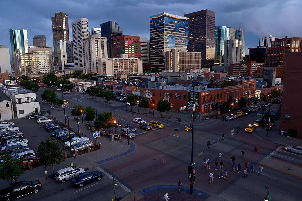 Denver skyscrapers beneath a blue sky, with cars and pedestrians in the foreground.