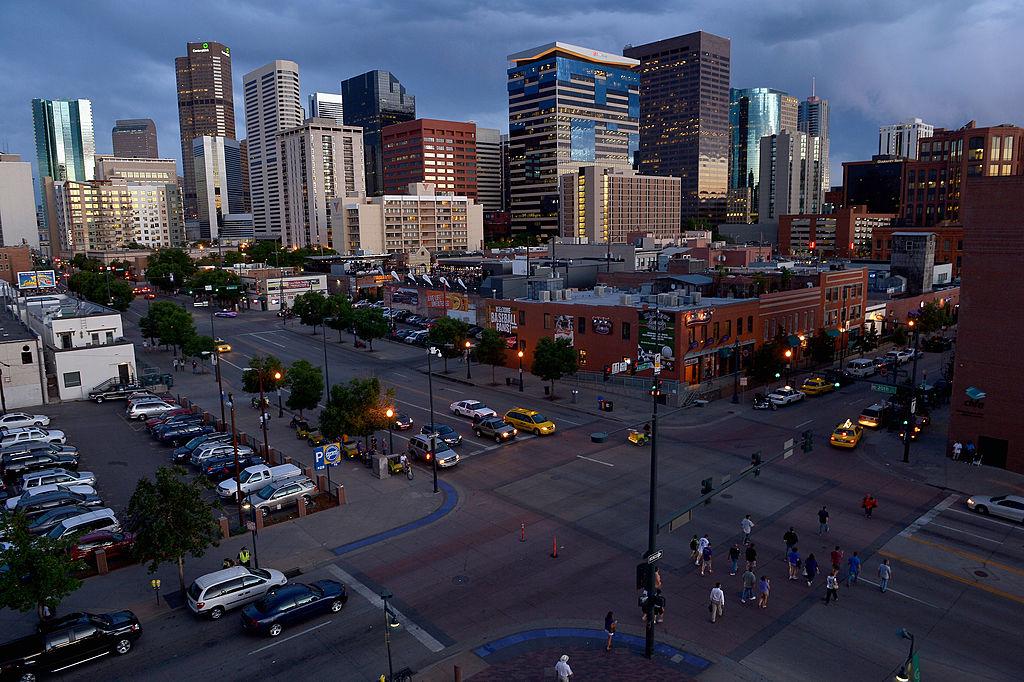 Denver skyscrapers beneath a blue sky, with cars and pedestrians in the foreground.