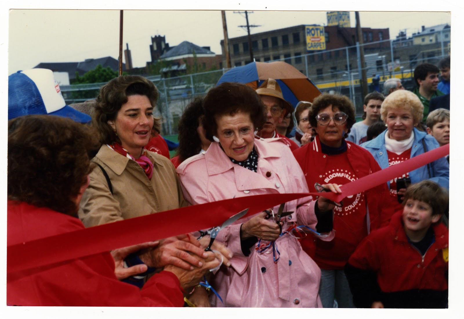 A woman in a long pink coat cuts a red ribbon with a large pair of scissors.