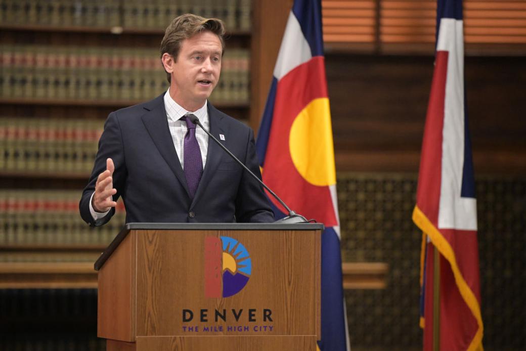 Mayor Mike Johnston speaks into a microphone at a podium that says “Denver The Mile High City.” 