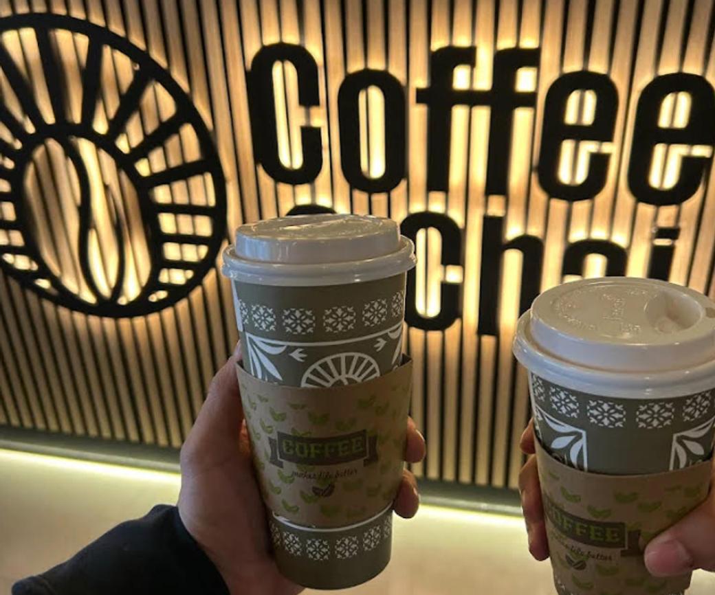 Hands holding coffee cups in front of Coffee & Chai sign.