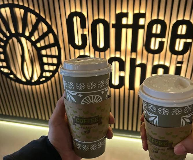 Hands holding coffee cups in front of Coffee & Chai sign.
