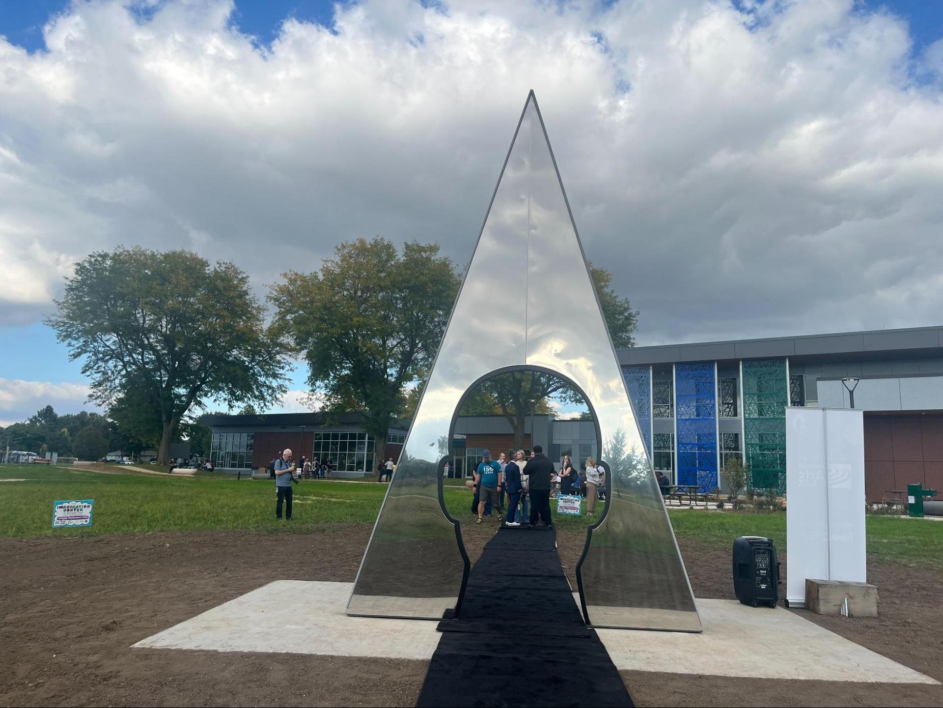 A triangular mirror statue sits on the grounds of the Imagination Center under cloudy skies.