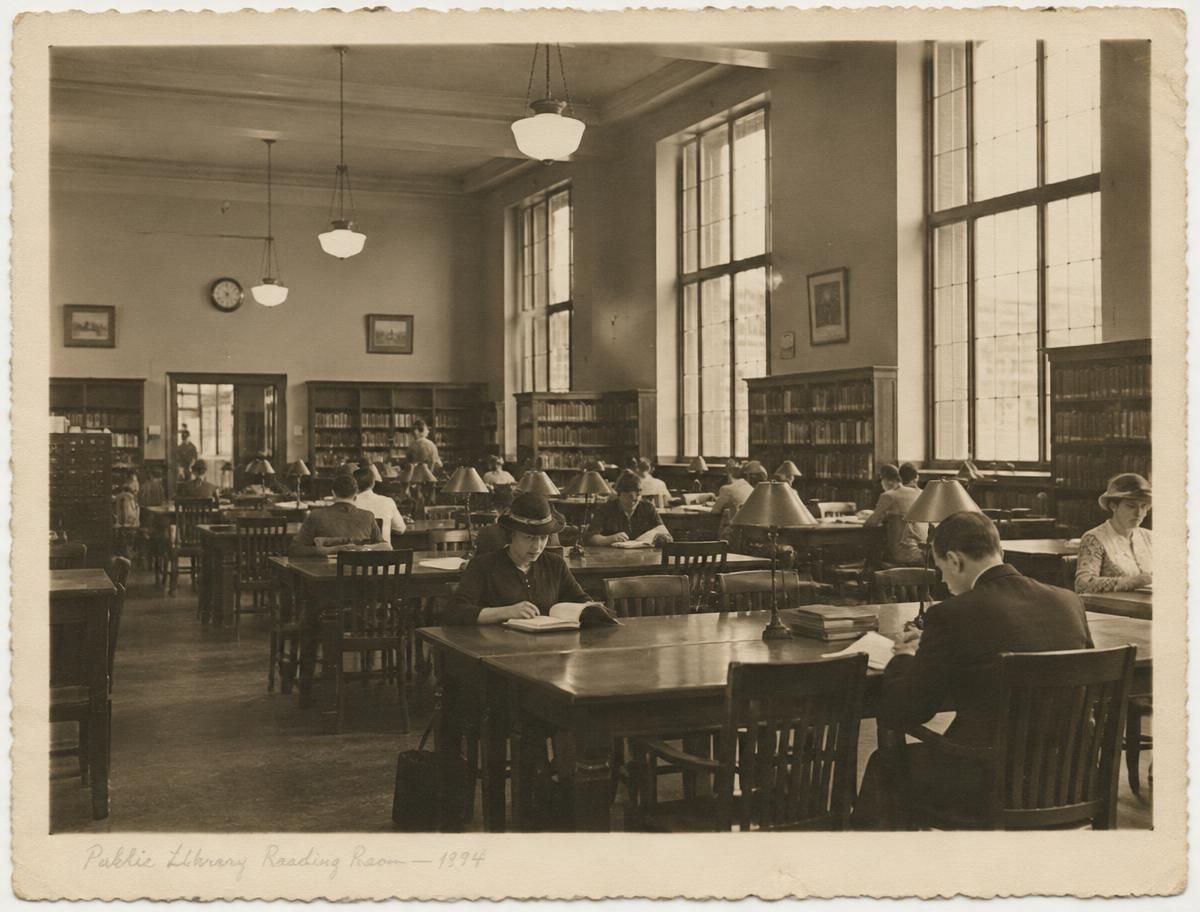 Sepia archival print of a busy public library reading room with desk lamps and tall windows