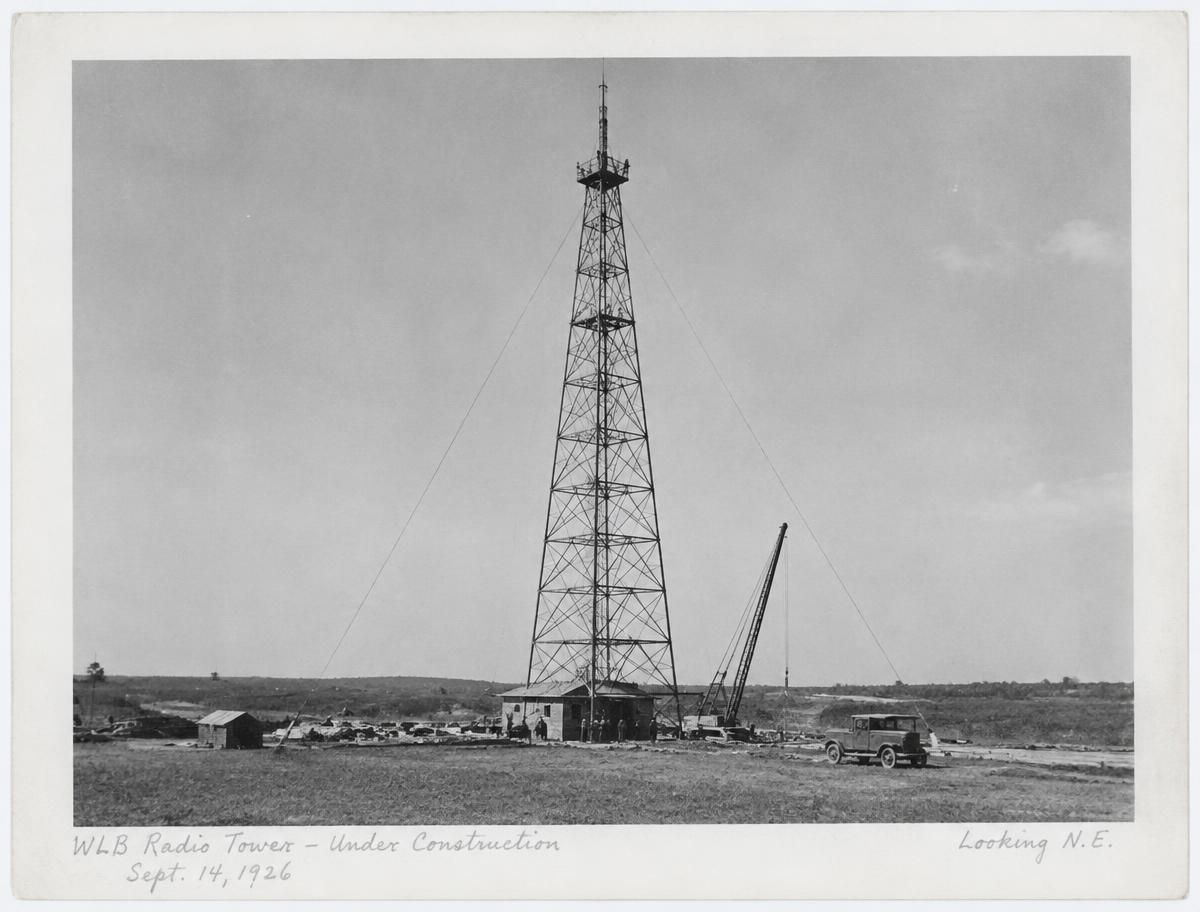Archival print of a lattice radio tower being built in an open field, with a handwritten caption in the print margin